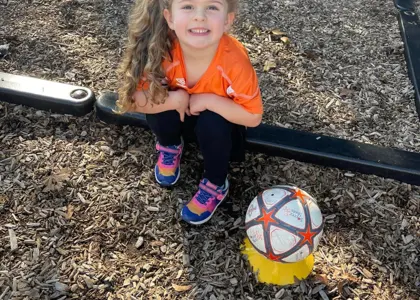 A smiling child sitting outside next to a soccer ball